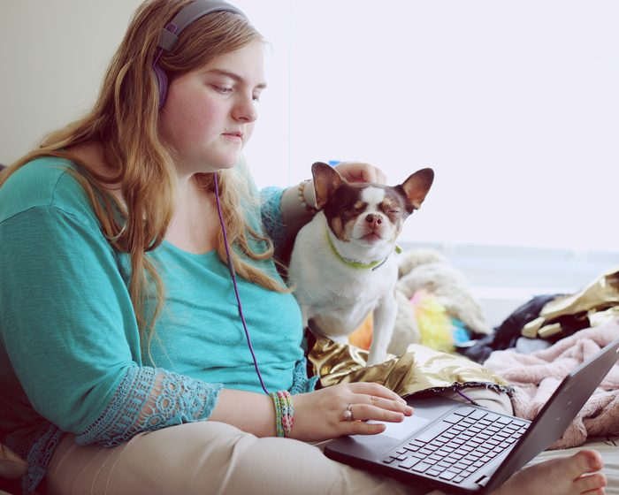 A young woman at home on her bed with a laptop and her dog.